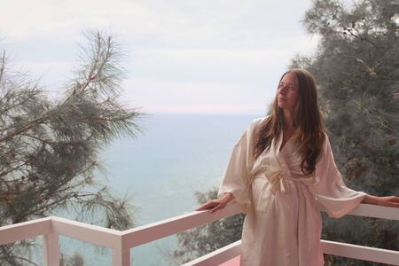 the concept of tourism, recreation-a beautiful girl with long hair, standing in a white coat on the balcony of the hotel overlooking the seaの写真素材
