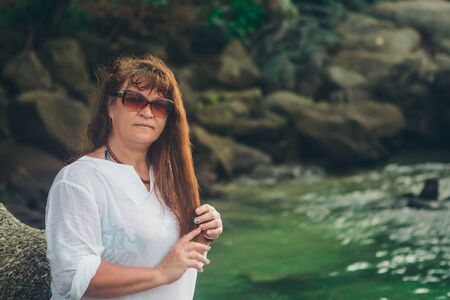 portrait of a tourist with long hair in sunglasses and a white t-shirt against the rocks and the sea. woman in travelの写真素材
