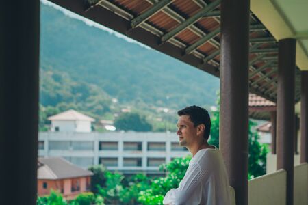 the concept of tourism, leisure is a beautiful man, basking in the white dress on the balcony of the hotel with views of tropical plants and a small buildingの写真素材