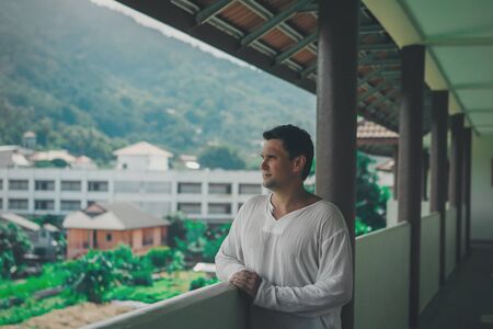 the concept of tourism, leisure is a beautiful man, basking in the white dress on the balcony of the hotel with views of tropical plants and a small buildingの写真素材