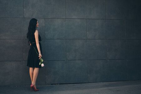 a beautiful brunette in a black dress and red shoes with a rose in her hands. Woman advertises accessories, clothes, shoesの写真素材