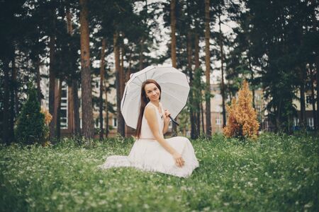 a beautiful brunette in a white dress with an umbrella sits on the grass. Beautiful girl in natureの写真素材