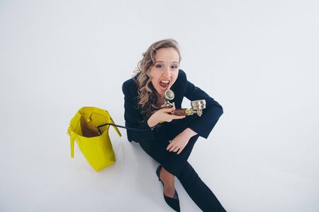 a businesswoman in a black and yellow suit pulls out of a yellow bag phone and yells at him. Beautiful woman sitting on floor on white background. the concept of the work in the office, business plan, fashionの写真素材
