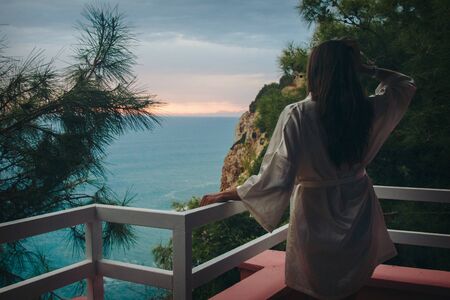 the concept of tourism, recreation-a beautiful girl with long hair, standing in a white coat on the balcony of the hotel overlooking the sea and the sunsetの写真素材