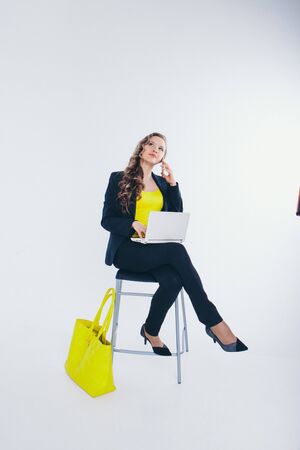 office concept, business plan, fashion-businesswoman in black and yellow suit talking on the phone and working on a laptop. big yellow bag. Beautiful woman sitting on chair on white backgroundの写真素材