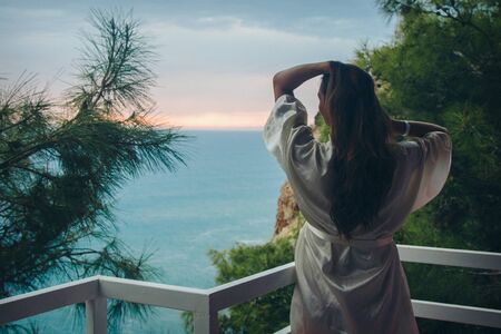 the concept of tourism, recreation-a beautiful girl with long hair, standing in a white coat on the balcony of the hotel overlooking the sea and the sunsetの写真素材