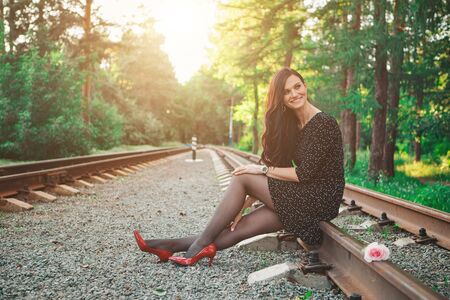 a beautiful brunette in a black dress and red shoes sits on the rails. Woman advertises accessories, cosmetics, makeup, clothesの写真素材