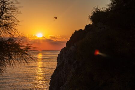 A skydiver flies with a colorful parachute tied to a speedboat on the sea against the backdrop of mountains and sunset. The concept of summer holidays, vacation, tourism.の写真素材