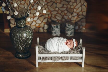 the baby is lying in a wooden bed under a blanket against the background of stacked firewood and a decorative vase. portrait of a small child. concept of childhood, health, IVF, children's furniture, interior, wood productsの写真素材