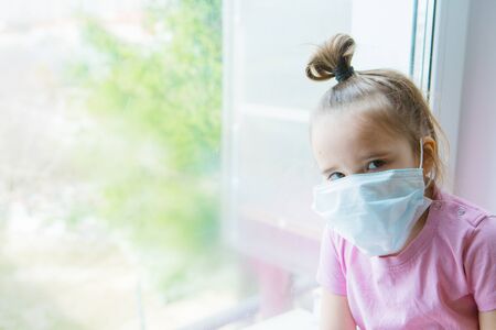 the girl in the medical mask leaned against the window. the girl looked out the window. a person is bored in self-isolation. I look at the world through glass. sad faceの写真素材