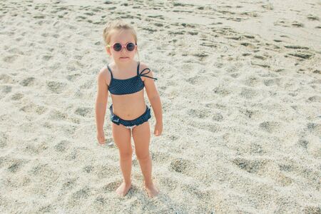 a small child walks on the beach. girl in a blue swimsuit and round glasses on a tripの写真素材