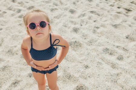 a small child walks on the beach. girl in a blue swimsuit and round glasses on a tripの写真素材