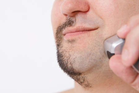 isolated on a white background: a man shaves his stubble. the guy cleans his beard with an electric razor. morning treatments in the bathroom. half of the beard close-up. copy spaceの写真素材