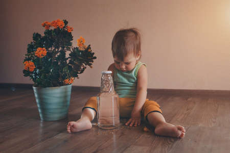 a small child is sitting on the floor next to a large flower in a pot and buttle with water. girl rips off a plantの写真素材
