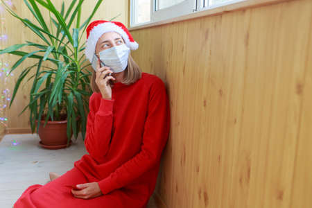 Celebration in self-isolation. christmas online congratulations: a woman in red Santa hats and medical mask congratulating family or friends with a tablet. Winter holidaysの写真素材