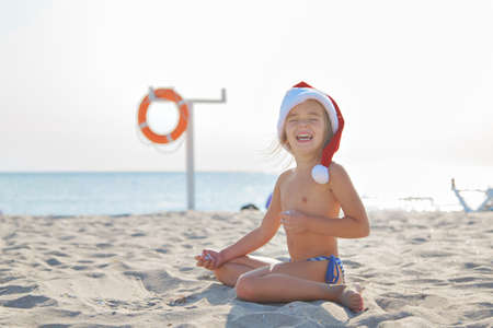 the girl is sitting and playing on the sand. Red Santa hat and lifebuoy on the sea background. Tourism during the new year holidays. Christmas decor and security. Copy spaceの写真素材