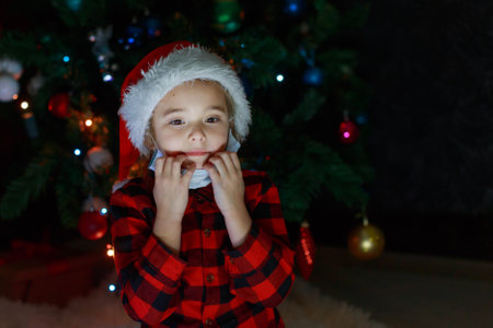 A little girl in a red Santa Hat and mask is sitting under the Christmas tree at home.の写真素材