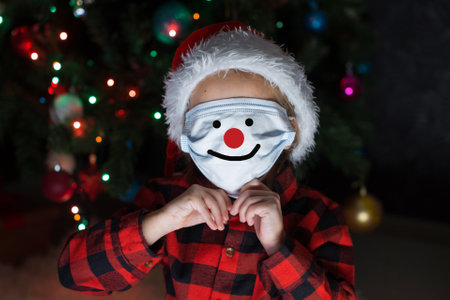 A little girl in a red Santa Hat and mask is sitting under the Christmas tree at home.の写真素材