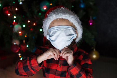 A little girl in a red Santa Hat and mask is sitting under the Christmas tree at home.の写真素材