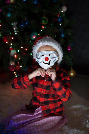 A little girl in a red Santa Hat and mask is sitting under the Christmas tree at home.の写真素材