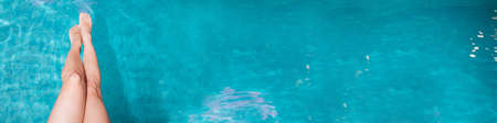 Close-up of slender female legs against the blue water by the pool on a hot summer day, under the sun. The girl dangles her legs in the water of the pool, resting, sunbathingの写真素材
