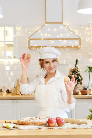 a cheerful chef prepares a Christmas cake. Lady cook in a cute apron breaking an egg into flour to...