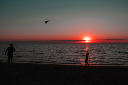 Dad and daughter fly a kite, their silhouettes can be seen against the background of the sun and the ocean. happy girl running with a kite at sunset outdoors. A child runs with a kite at sunset against the backdrop of the sea. Family vacation. High quality photo.の写真素材