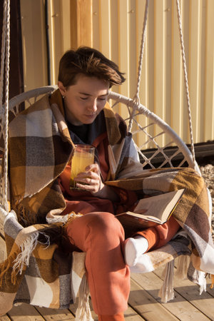 A woman with a short haircut wrapped herself in a plaid blanket and drinks an energy drink. The girl drinks water in the morning on the balcony and the sun shines on her. Healthy food, vitamins.の写真素材