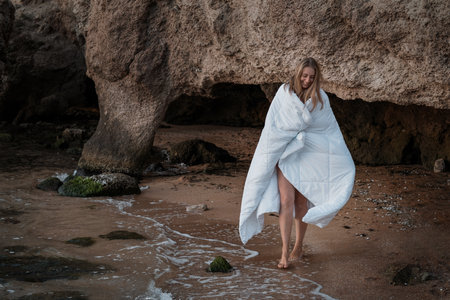 a girl in a white blanket walks along the beach at the sea, near the rocks. Beautiful, slender legs walk along the beach. Cream for varicose veins. High quality photoの写真素材