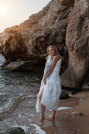 a girl in a white blanket walks along the beach at the sea, near the rocks. Beautiful, slender legs walk along the beach. Cream for varicose veins. High quality photoの写真素材