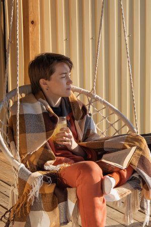 A woman with a short haircut wrapped herself in a plaid blanket and drinks an energy drink. The girl drinks water in the morning on the balcony and the sun shines on her. Healthy food, vitamins.の写真素材
