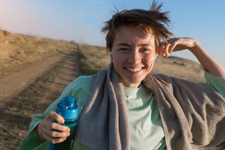 a girl with short brown hair runs along the road and smiles with a towel and a bottle of water. Sports girl doing exercises in the morning. Healthy back, self-love, joy and relaxation. High quality photoの写真素材