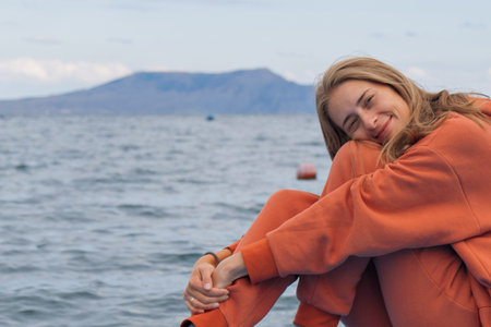 A woman tourist looks at the mountains and the sea in an orange sports suit, jacket and backpack....