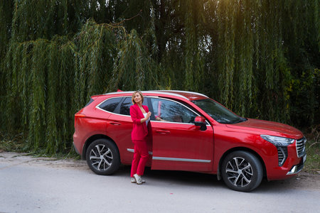A woman in a red Chinese car drinks coffee in a business suit. Online work in nature. Freelance, travel, remote work. Freedom of movement, model in the forest. High quality photoの写真素材