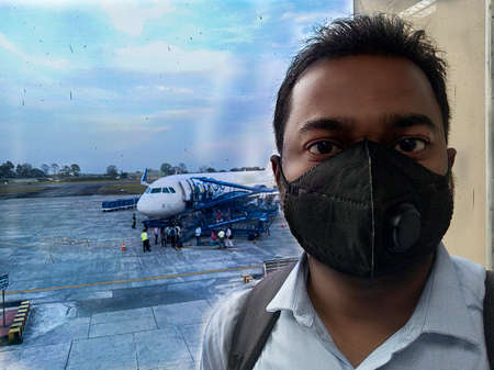 Smart Asian man wearing face mask protection in prevention for Corona virus or Covid-19 and micro dust pm 2.5 in air while standing at waiting area at Airport terminal and behind passengers are boarding an aeroplane.のeditorial素材