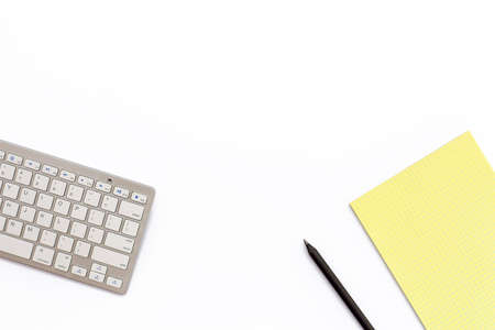 work office desk with a keyboard, a yellow notepad and a black pencil on white background. Minimal business concept. Flat lay. Top view.の写真素材