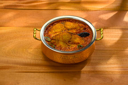 Seer Fish curry, traditional Indian fish curry ,kerala special ,arranged in a copper serving bowl garnished with malabar tamarind and fresh green chilli on a wooden textured with abstract shadow on background,isolated.の写真素材