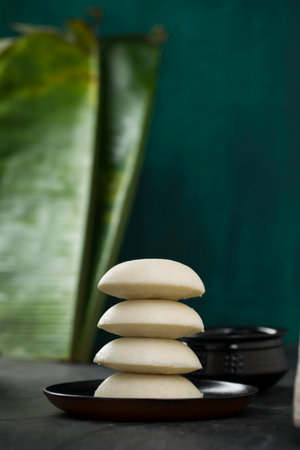Idly or Idli, south indian main breakfast item which is placed in a heap one over other beautifully arranged in a black plate  on dark green  texture with kitchen  background, selective focus.の写真素材