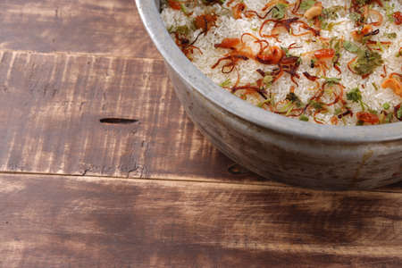 Dum chicken biriyani, close up image of Kerala-Thalassery biriyani garnished with fried onion,cashew nut,raisens and coriander leaf which is arranged in a big aluminium cooking pot which is placed on wooden textured background.の写真素材