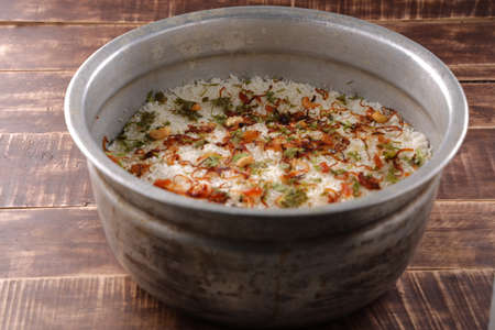 Dum chicken biriyani, close up image of Kerala-Thalassery biriyani garnished with fried onion,cashew nut,raisens and coriander leaf which is arranged in a big aluminium cooking pot which is placed on wooden textured background.の写真素材