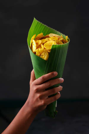 Dried banana chips or banana waffers, a hand holding banana leaf cone filled with waffers, grey...