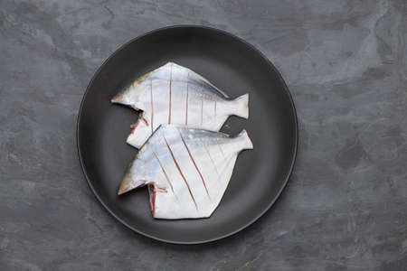 Raw white pomfret, ready to cook pomfret arranged in a black plate with grey colour background'の写真素材