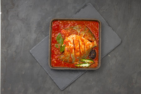 White pomfret masala curry , made in south indian style with thick red gravy and garnished with green chilli,curry leaves and malabar tamarind arranged in an iron sizzler with grey colour background.の写真素材