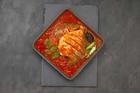 White pomfret masala curry , made in south indian style with thick red gravy and garnished with green chilli,curry leaves and malabar tamarind arranged in an iron sizzler with grey colour background.の写真素材