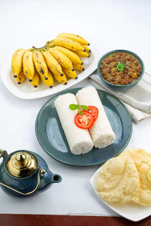 PUTTU -Kerala special breakfast made using rice flour which is arranged in an aquarustic plate with pieces of paapad, bowl of curry and banana placed besides it on a white background,の写真素材