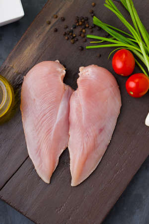 Fresh skinless raw chicken breastfillet arranged on a wooden board and garnished with tomato, spring onion and oil on grey textured background with package box nearbyの写真素材