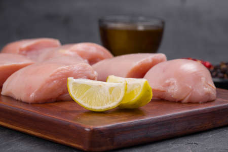 Raw chicken tender fry cut without skin arranged on wooden board and garnished with oil,lemon slices , chilly flakes and black pepper on stone textured or graphite colour backgroundの写真素材