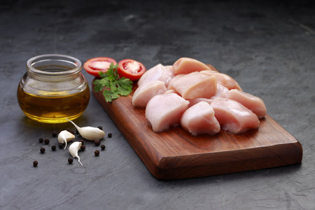Raw chicken tender fry cut without skin arranged on wooden board and garnished with oil,coriander leaf,tomato slices,garlic and pepper with  stone textured or graphite colour backgroundの写真素材
