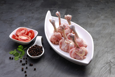 Raw chicken lollipop,five pieces of chicken lollipop arranged on a white serving tableware with  tomato slices  and pepper  on background with grey textured baseの写真素材