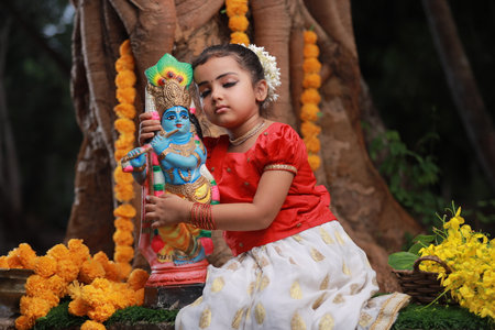 A cute small girl child wearing  Kerala dress-golden colour long skirt and red blouse, sitting under banyan tree with statue of lord krishna-golden shower flower near byの写真素材